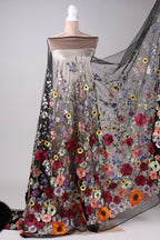 A mannequin wearing a long, flowing dress with a sheer overlay featuring a vibrant floral pattern in various colors, including red, orange, yellow, and purple.