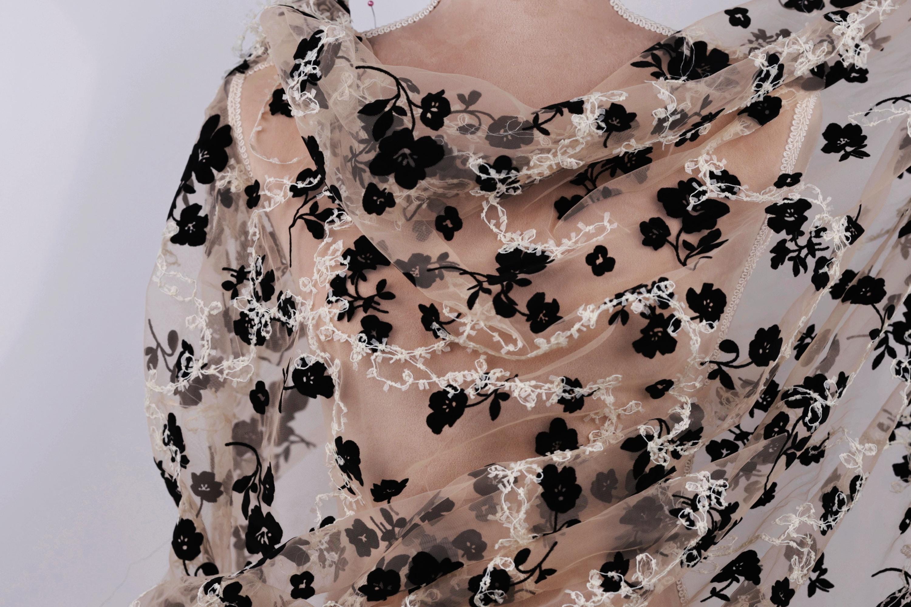 a close-up view of a delicate, sheer fabric with a floral pattern. The fabric appears to be made of sheer material, giving it a delicate and airy appearance. The floral pattern consists of black flowers interspersed with white flowers, creating a visually striking contrast against the light background. The fabric is draped over the back of a person, adding an elegant and feminine touch to the image.