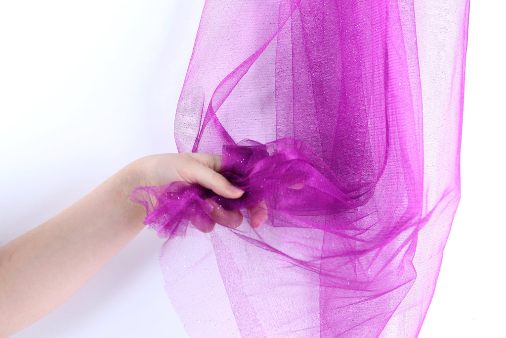 A hand is holding a piece of purple tulle fabric against a white background.
