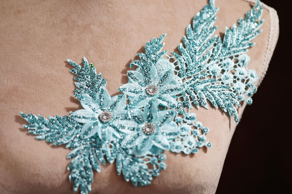 A close-up view of a delicate, intricately designed fabric with a floral pattern. The fabric appears to be made of a light-colored material, possibly lace or a similar type of material. The design features a combination of blue and green colors, with the flowers and leaves being the central focus of the image.