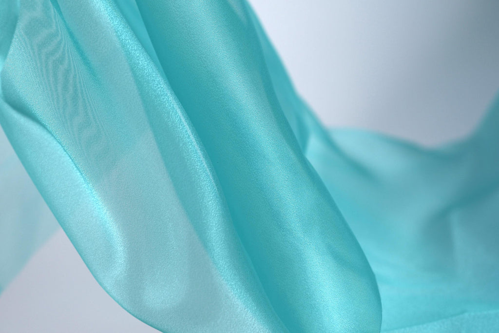 A close-up view of a flowing, turquoise-colored fabric, with a blurred background that emphasizes the fabric's texture and movement.