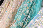 a close-up view of a variety of colorful, intricately embroidered fabric pieces. The fabric appears to be a blend of pastel colors, with floral and leaf patterns, and is adorned with sequins and beads. The fabric pieces are arranged in a way that showcases their unique designs and textures, creating a visually appealing display.