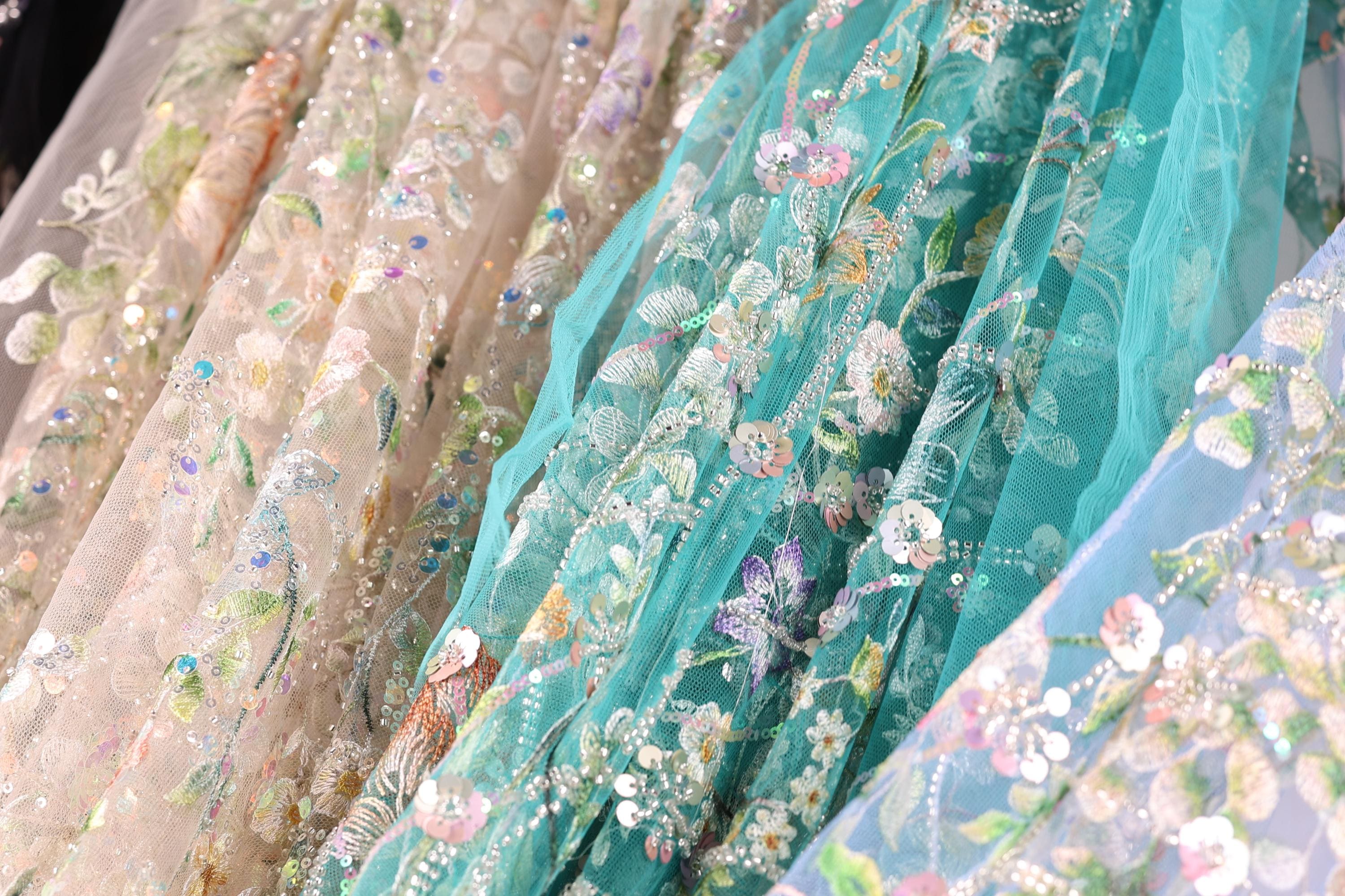 a close-up view of a variety of colorful, intricately embroidered fabric pieces. The fabric appears to be a blend of pastel colors, with floral and leaf patterns, and is adorned with sequins and beads. The fabric pieces are arranged in a way that showcases their unique designs and textures, creating a visually appealing display.