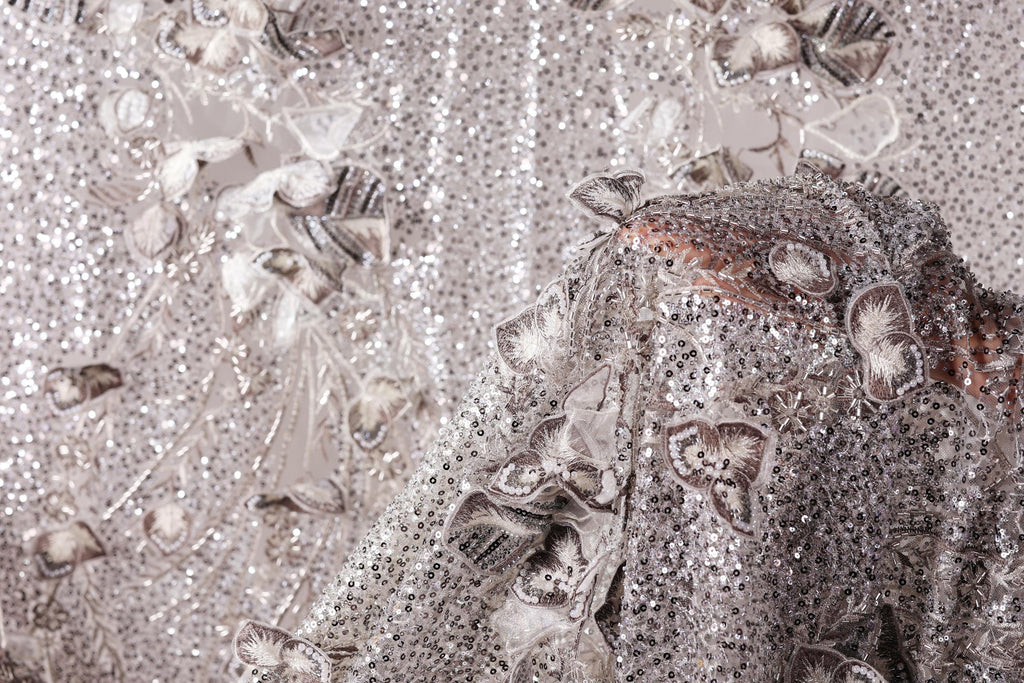 The image is a close-up of a fabric with a silver sequin pattern, featuring a variety of shapes and textures.