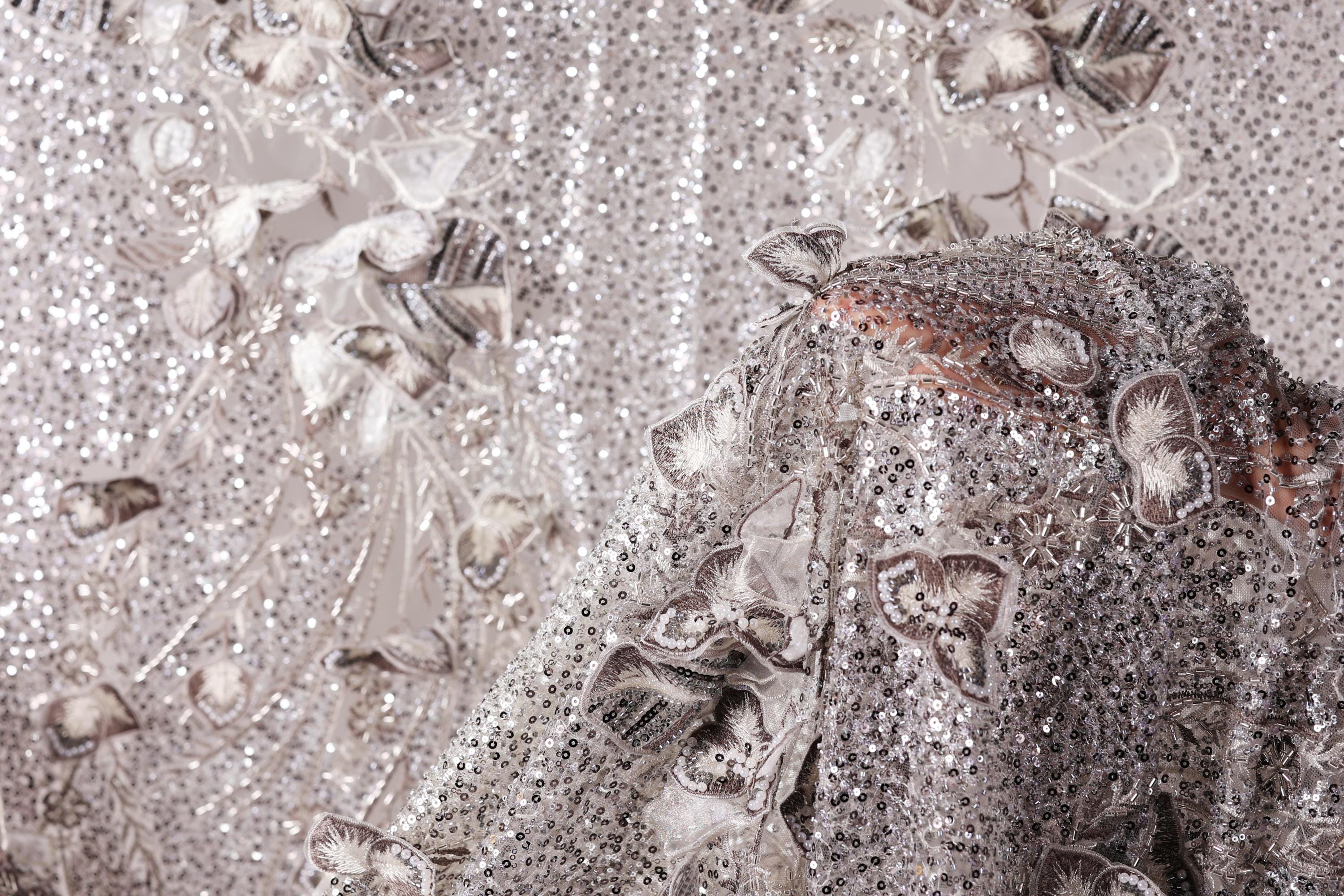The image is a close-up of a fabric with a silver sequin pattern, featuring a variety of shapes and textures.