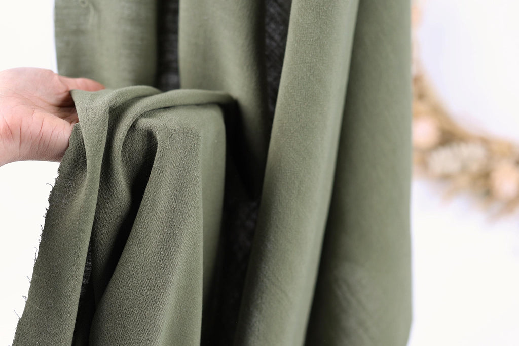 A hand is holding a piece of olive green fabric, which appears to be a type of cloth or textile.