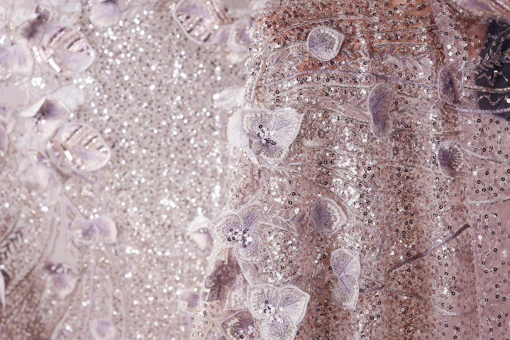 The image is a close-up of a fabric with a floral pattern, featuring small purple flowers and sequins. The fabric appears to be a light pink color, with a textured and shiny appearance.