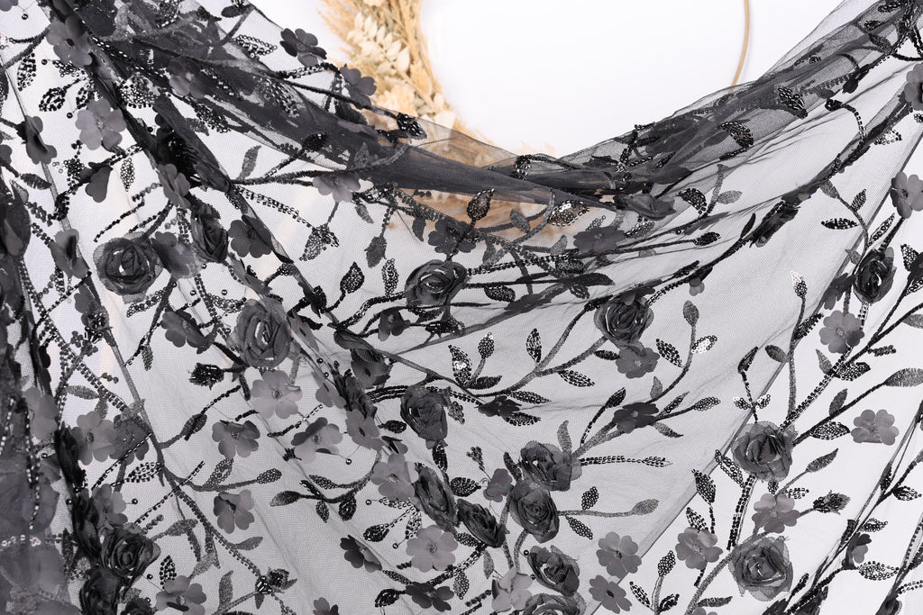 a close-up view of a black lace fabric with a floral pattern. The fabric appears to be made of sheer material, with a delicate and intricate design featuring black lace and floral elements. The fabric is displayed against a plain white background, highlighting its texture and design.