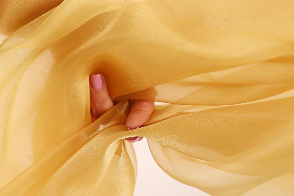 A hand is holding a piece of yellow fabric, which appears to be a flowing, sheer material. The hand is visible in the center of the image, with the fabric partially covering it.