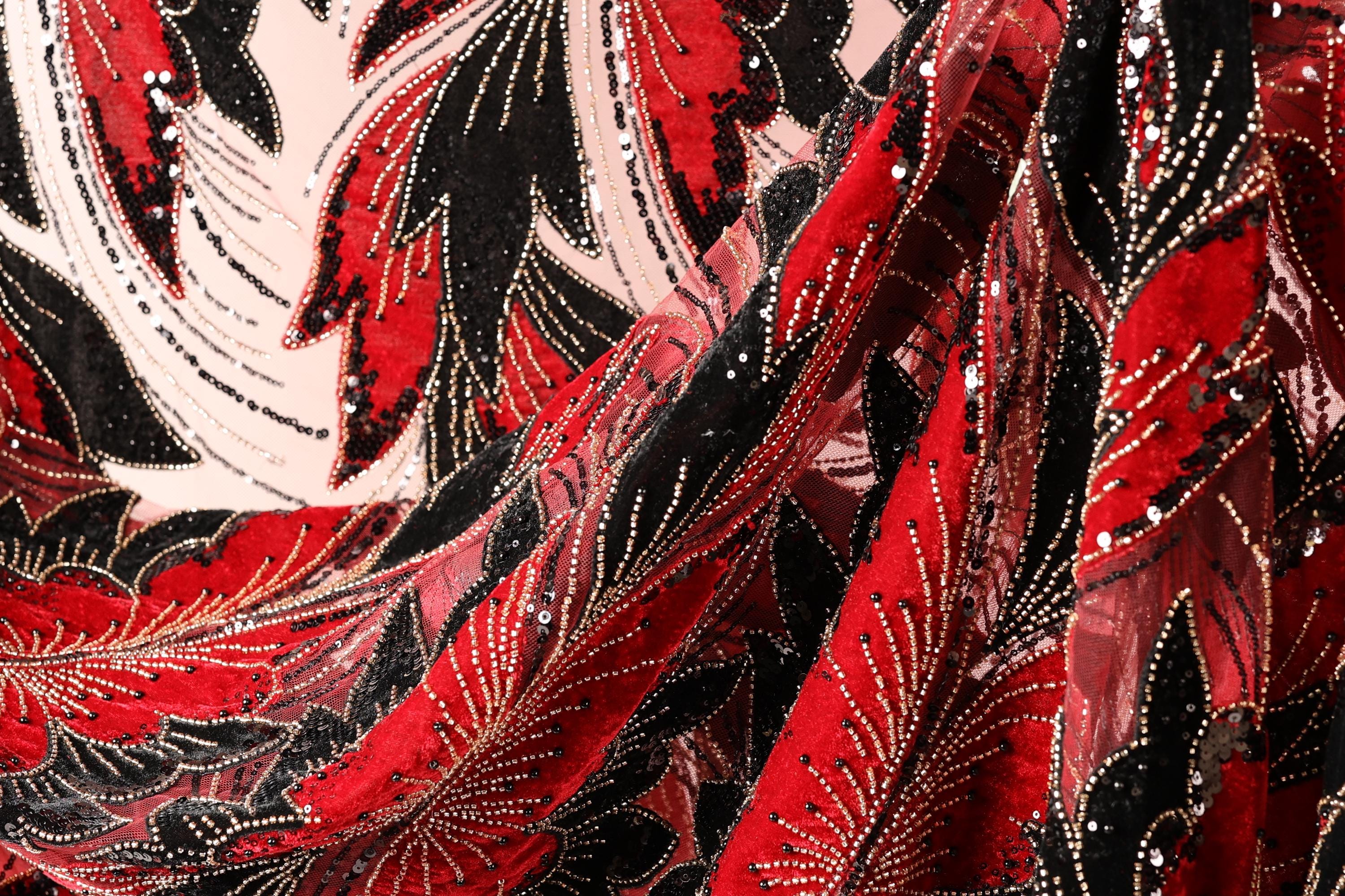 The image is a close-up of a fabric with a striking red and black pattern, featuring a repeating floral design with red leaves and black stems. The fabric appears to be made of a shiny, sequined material, adding a touch of sparkle and elegance to the design.