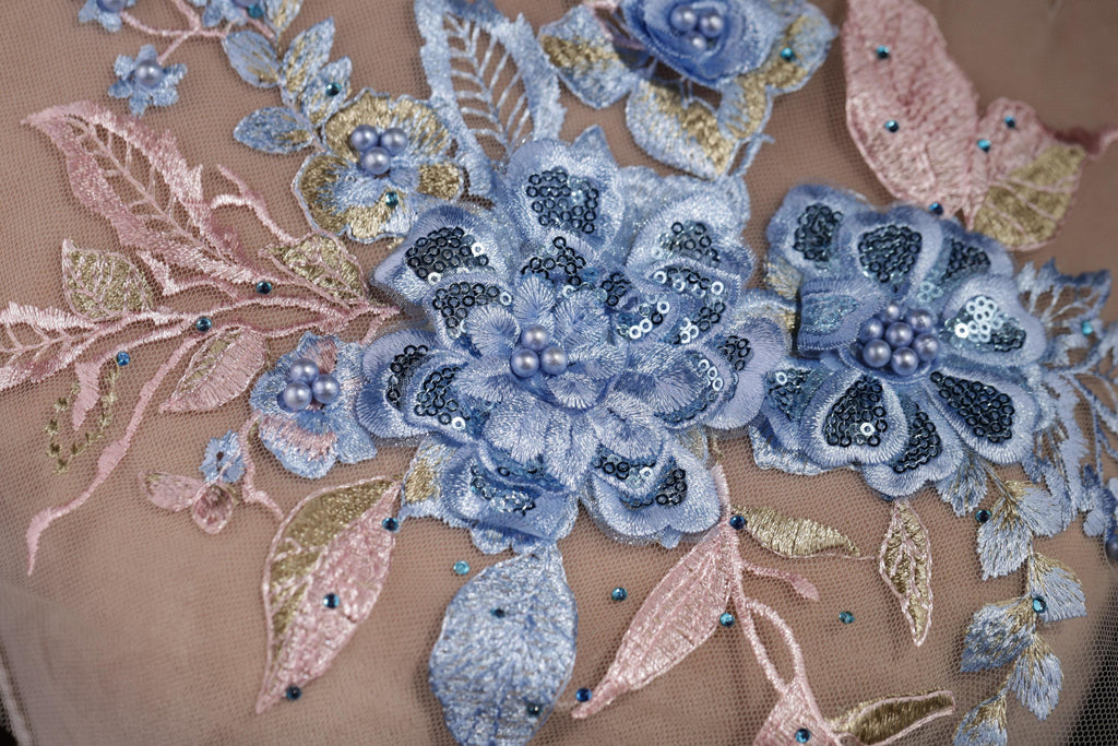 a close-up view of a delicate, intricately embroidered floral design on a fabric surface. The design is composed of various blue and pink flowers, leaves, and sequins, creating a visually appealing and detailed pattern. The flowers are the central focus of the image, with the leaves and sequins adding depth and texture to the overall composition.