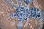 a close-up view of a delicate, intricately embroidered floral design on a fabric surface. The design is composed of various blue and pink flowers, leaves, and sequins, creating a visually appealing and detailed pattern. The flowers are the central focus of the image, with the leaves and sequins adding depth and texture to the overall composition.