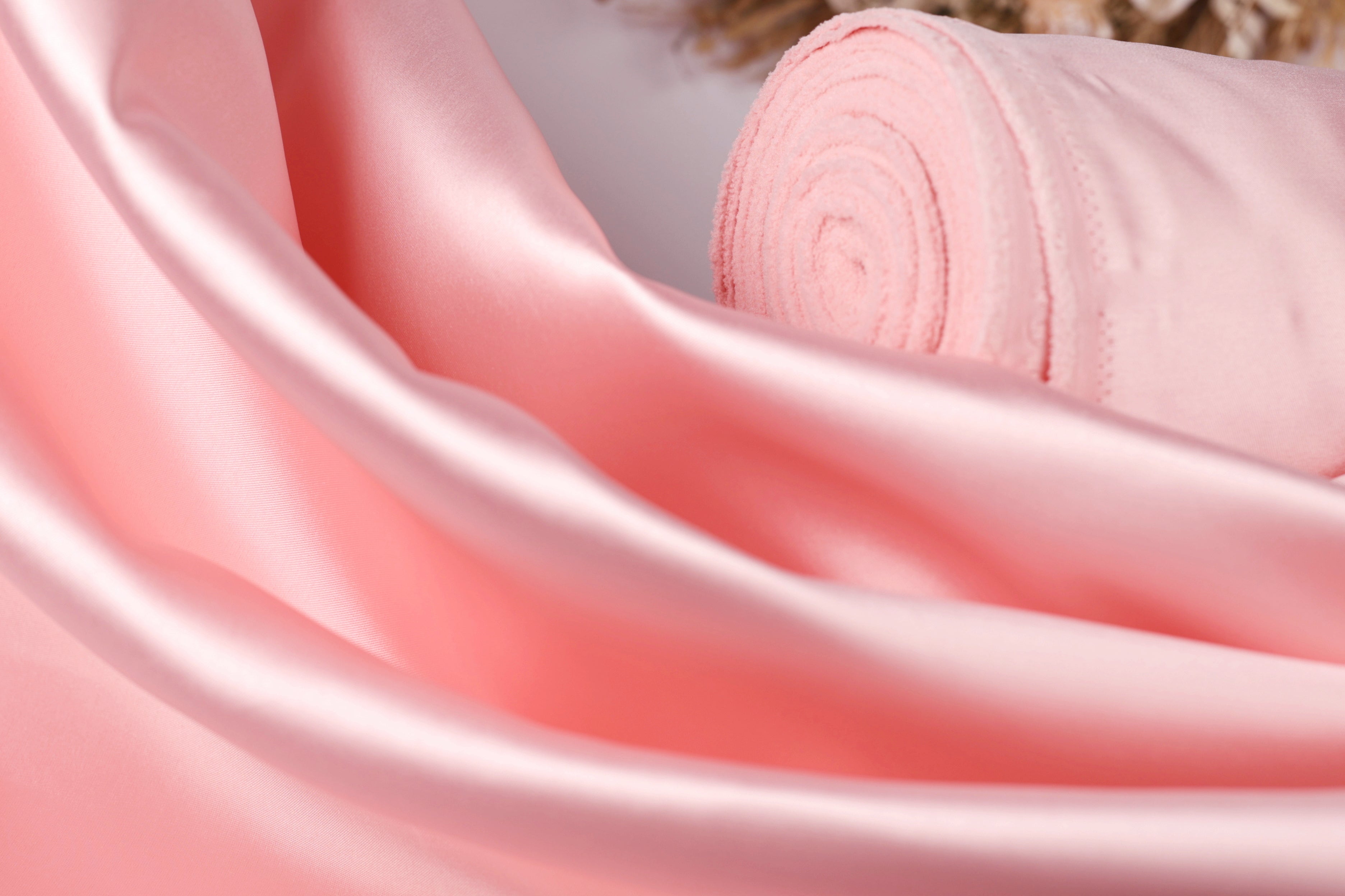 A close-up view of a soft, pink satin fabric, with a rolled-up piece of the fabric visible in the background.