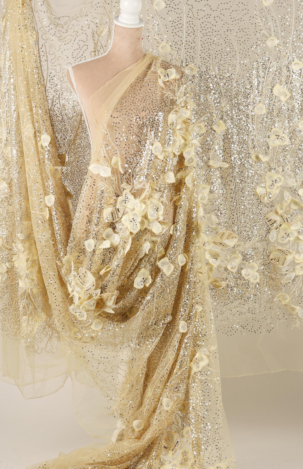 a mannequin wearing a gold sequin dress with a sheer, flower-like design. The dress has a long, flowing skirt and a sheer overlay, creating a shimmering effect. The dress appears to be made of sheer fabric, which adds to its delicate and elegant appearance.