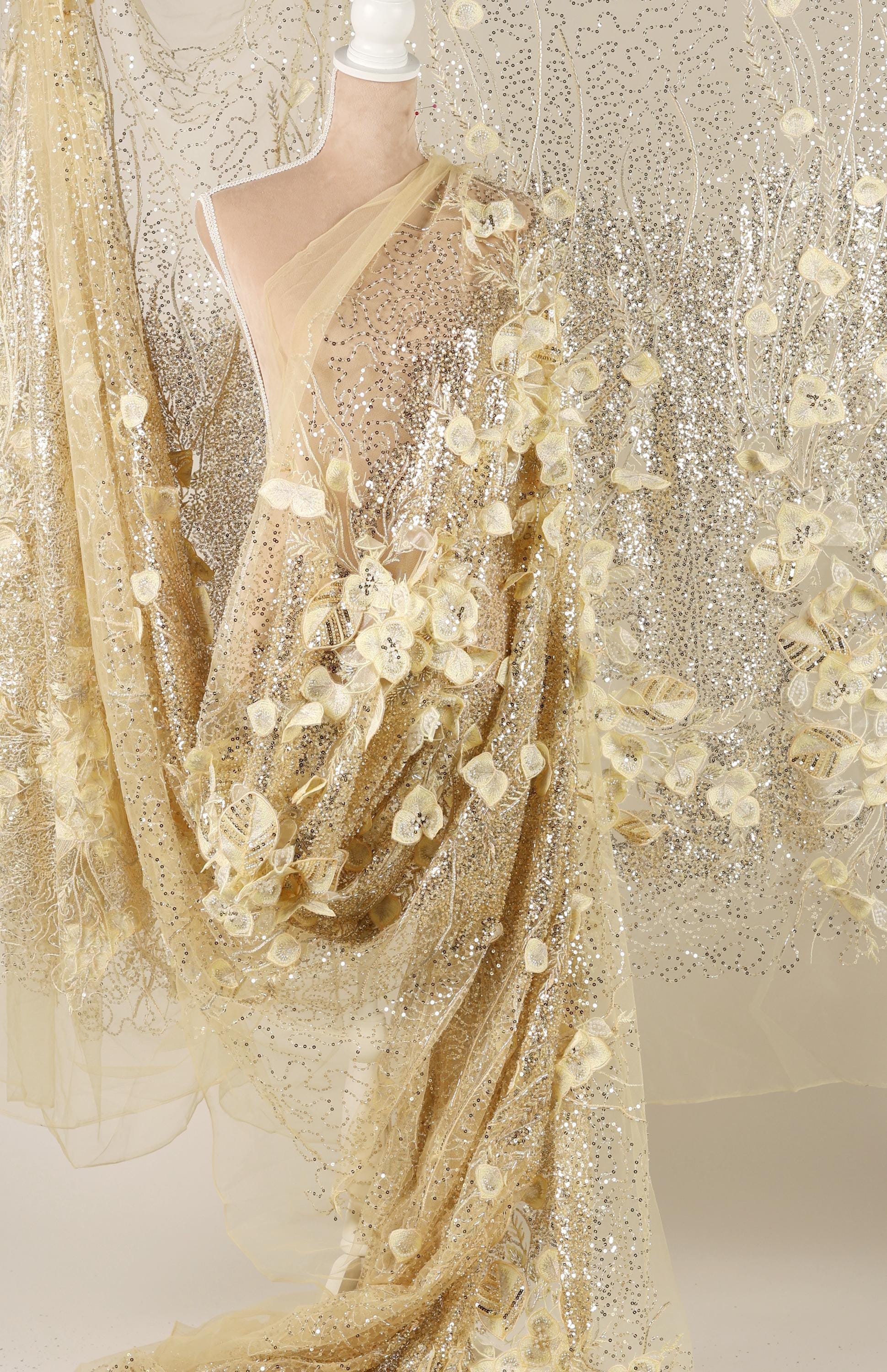 a mannequin wearing a gold sequin dress with a sheer, flower-like design. The dress has a long, flowing skirt and a sheer overlay, creating a shimmering effect. The dress appears to be made of sheer fabric, which adds to its delicate and elegant appearance.