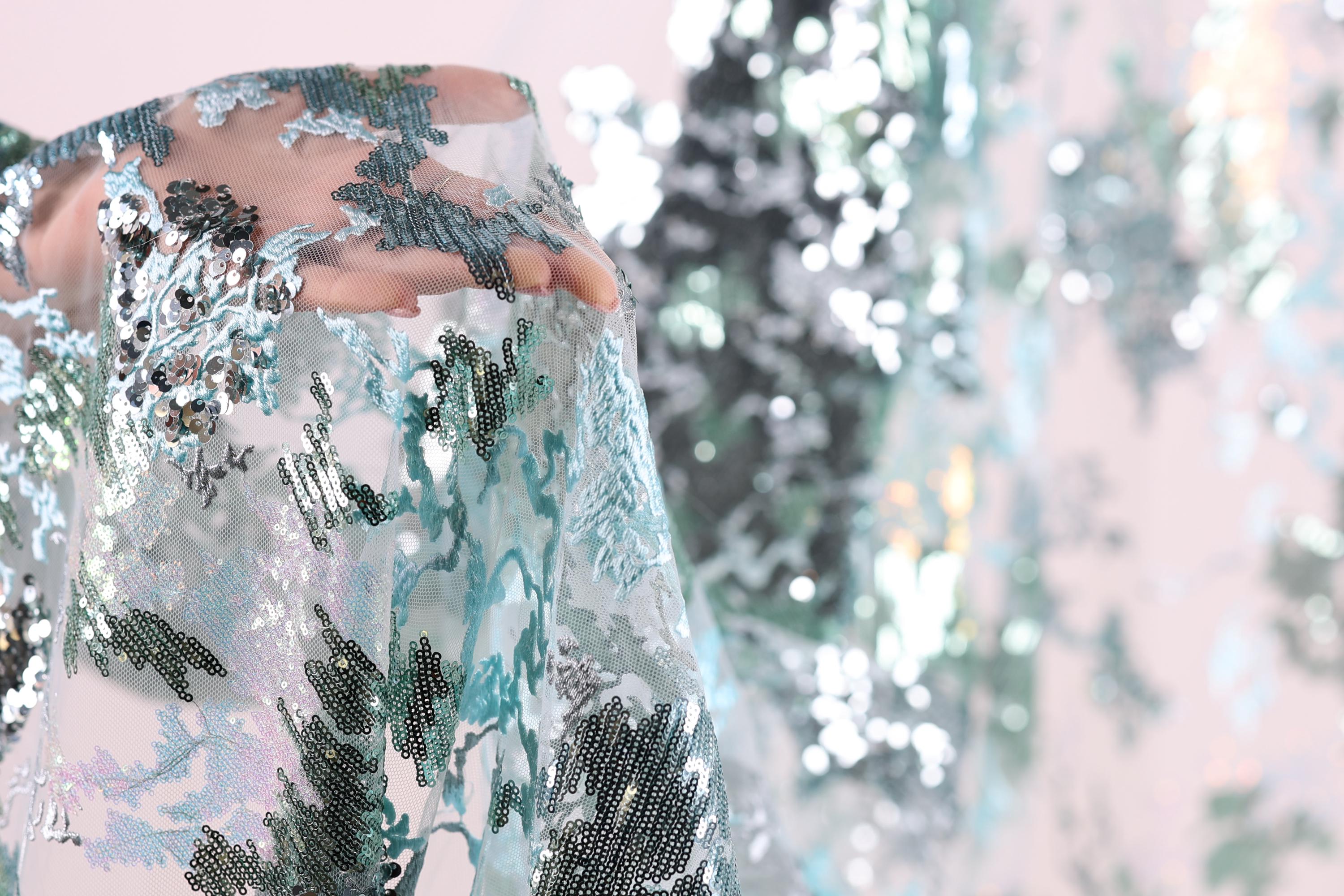 A close-up view of a hand holding a piece of fabric with a floral pattern, featuring a mix of blue, green, and silver sequins.