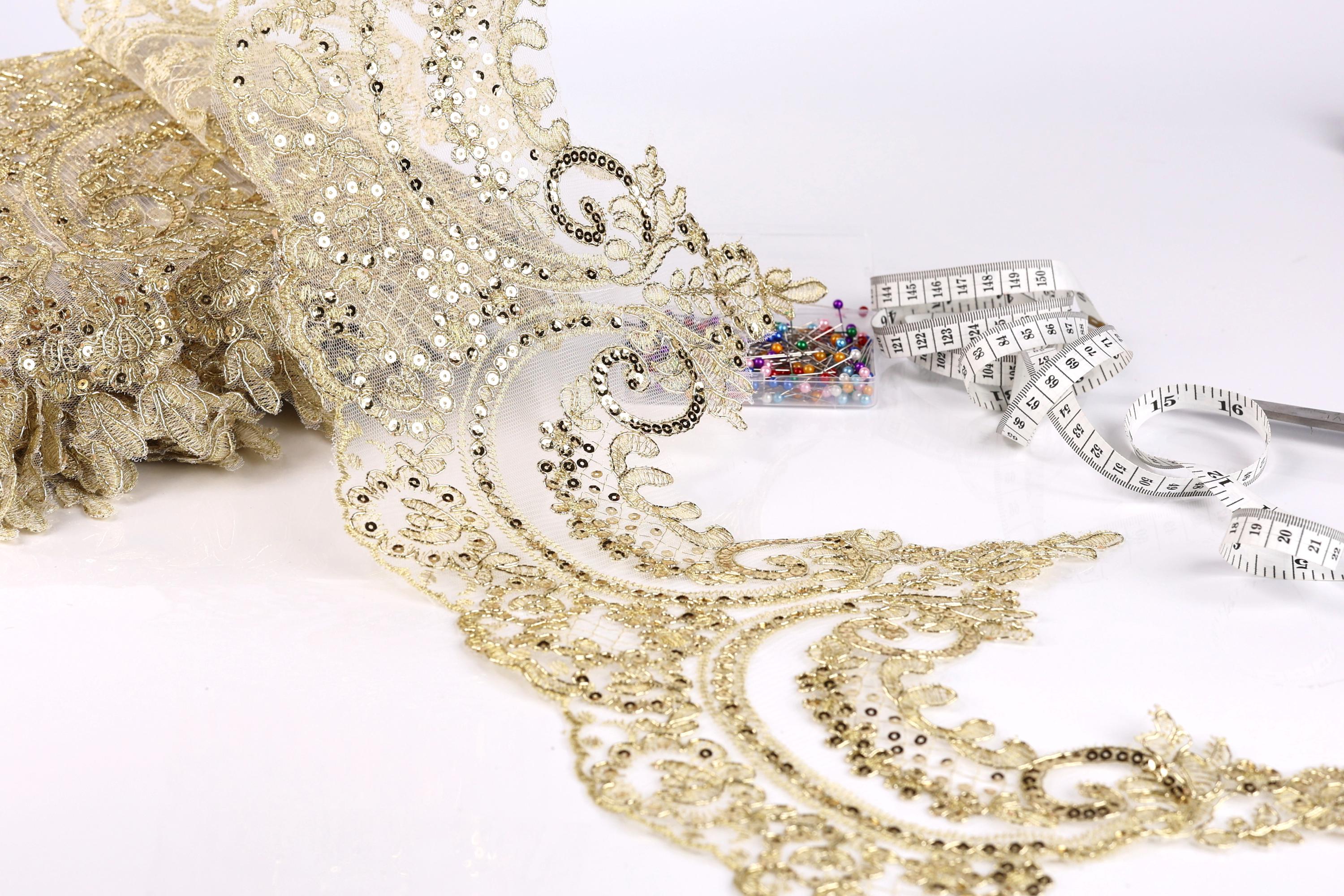 a close-up view of a gold sequin lace fabric with a measuring tape and a small box of sewing needles.