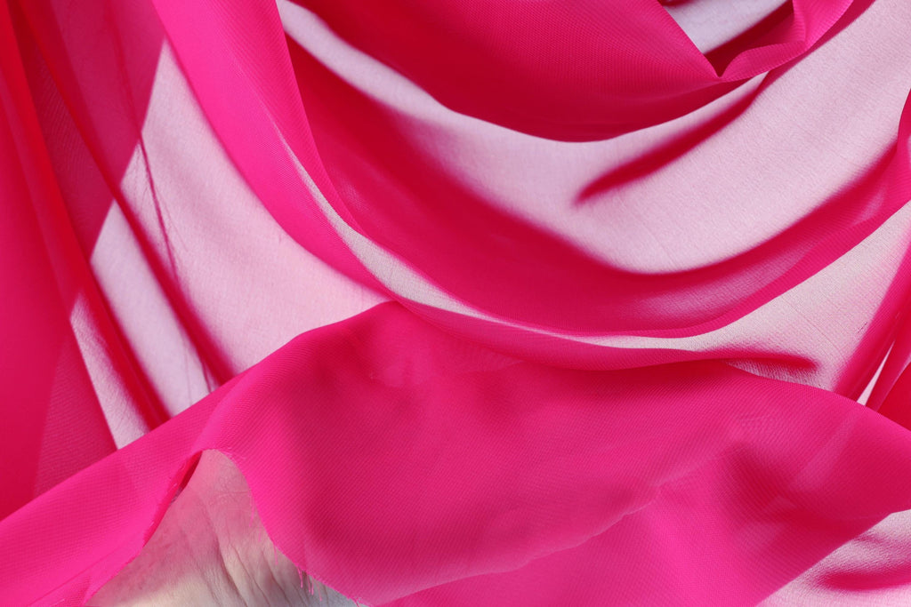 The image is a close-up of a vibrant pink fabric, with a soft, flowing texture. The fabric appears to be made of silk or chiffon, and it is slightly wrinkled, giving it a delicate and elegant appearance. The background is a light pink color, which complements the pink fabric and creates a harmonious color palette.