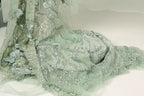 a close-up view of a delicate, intricately designed fabric with a floral pattern. The fabric appears to be made of a light green or mint-colored material, with a shimmering, sequined effect that adds a touch of elegance and sophistication to the design. The fabric is spread out on a surface, allowing for a detailed examination of its texture and patterns.