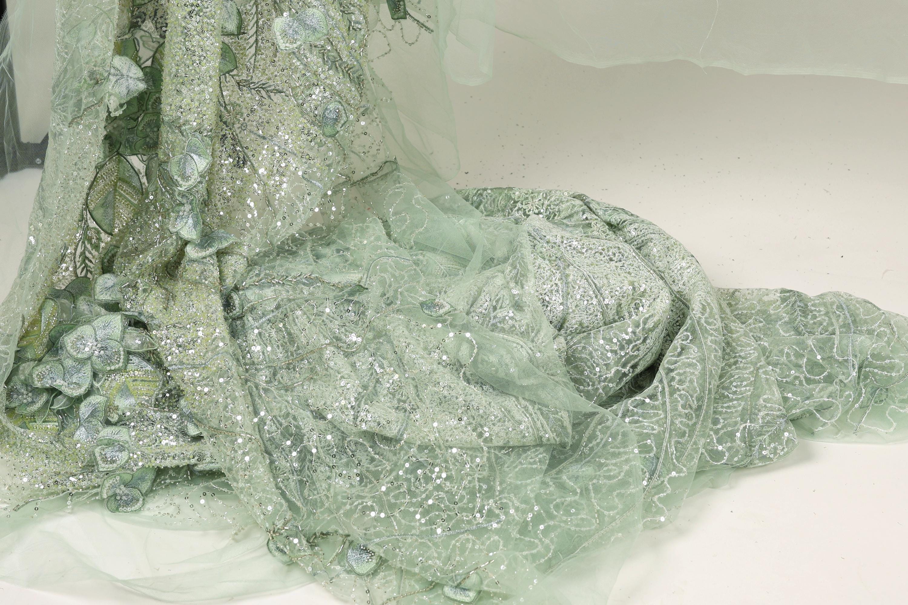 a close-up view of a delicate, intricately designed fabric with a floral pattern. The fabric appears to be made of a light green or mint-colored material, with a shimmering, sequined effect that adds a touch of elegance and sophistication to the design. The fabric is spread out on a surface, allowing for a detailed examination of its texture and patterns.