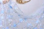 a delicate, light blue fabric with a floral pattern, including blue flowers and leaves. The fabric appears to be a sheer or sheer lace material, with a soft and flowing texture. The background is a plain white wall, which allows the blue fabric to stand out prominently.