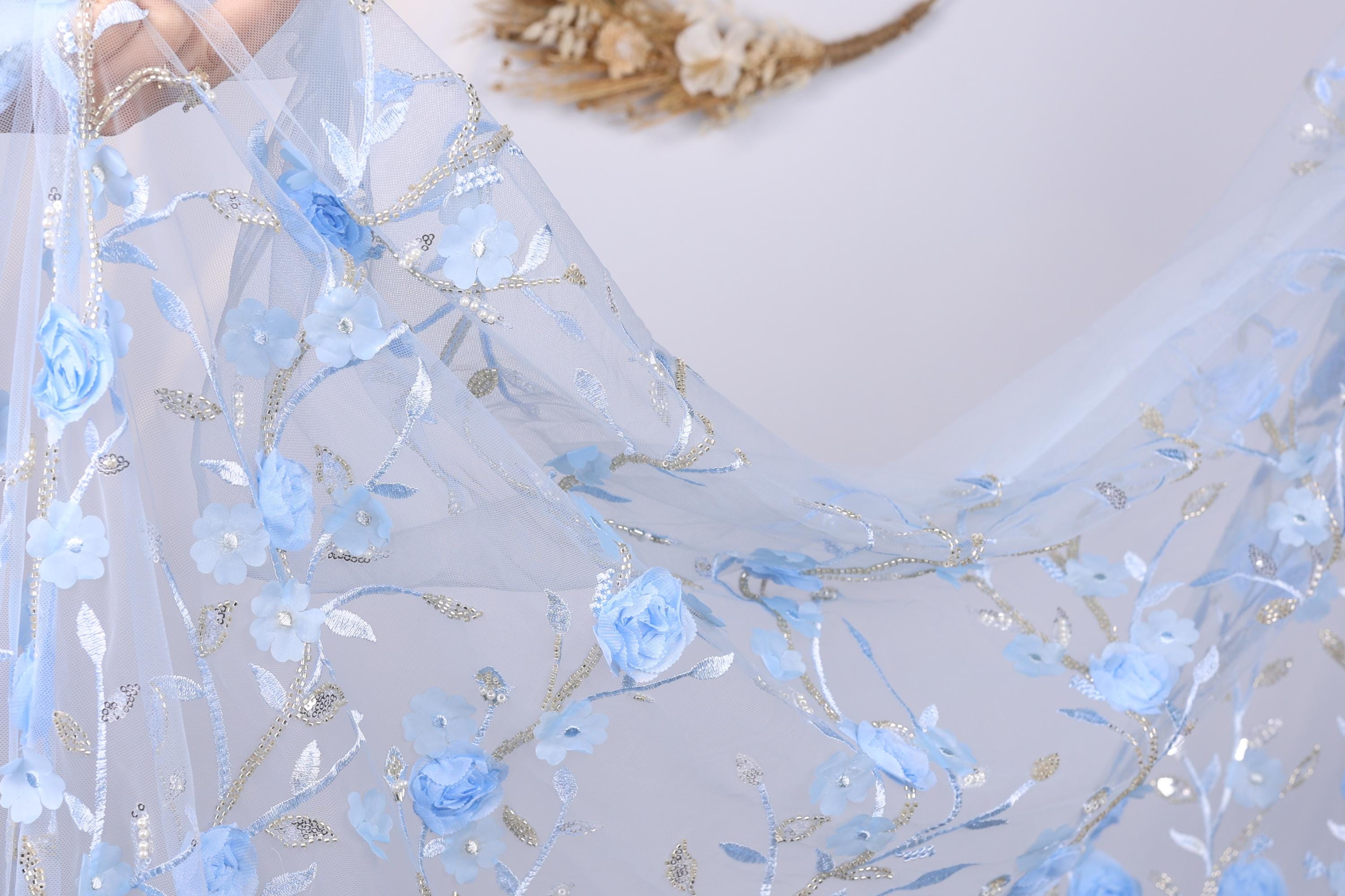 a delicate, light blue fabric with a floral pattern, including blue flowers and leaves. The fabric appears to be a sheer or sheer lace material, with a soft and flowing texture. The background is a plain white wall, which allows the blue fabric to stand out prominently.