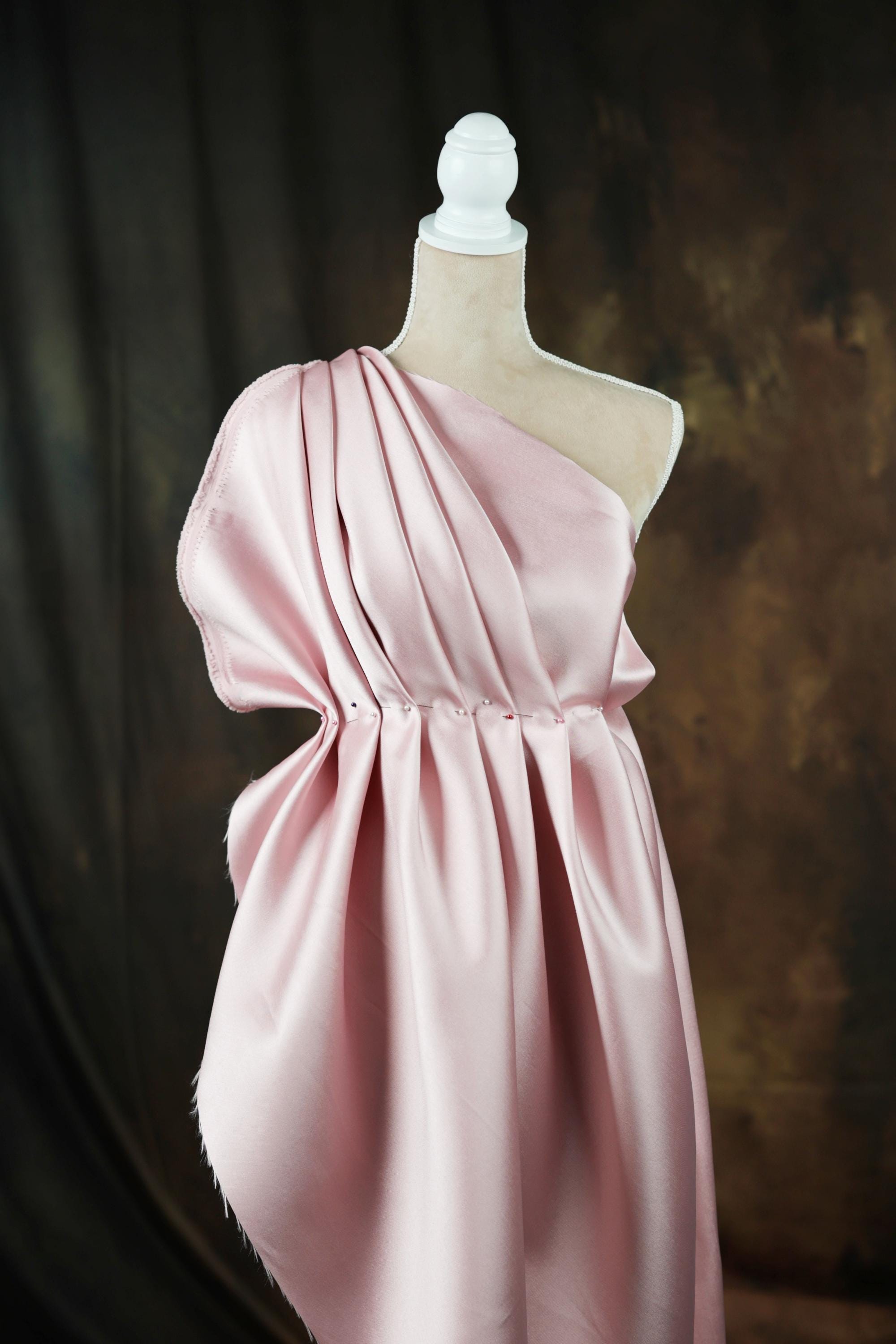A pink satin one-shoulder dress displayed on a mannequin against a dark background.