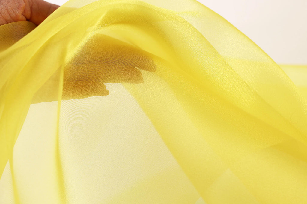 A hand is holding a yellow fabric, which appears to be a sheer material. The fabric is being held up against a white background, creating a contrast between the yellow and the white.
