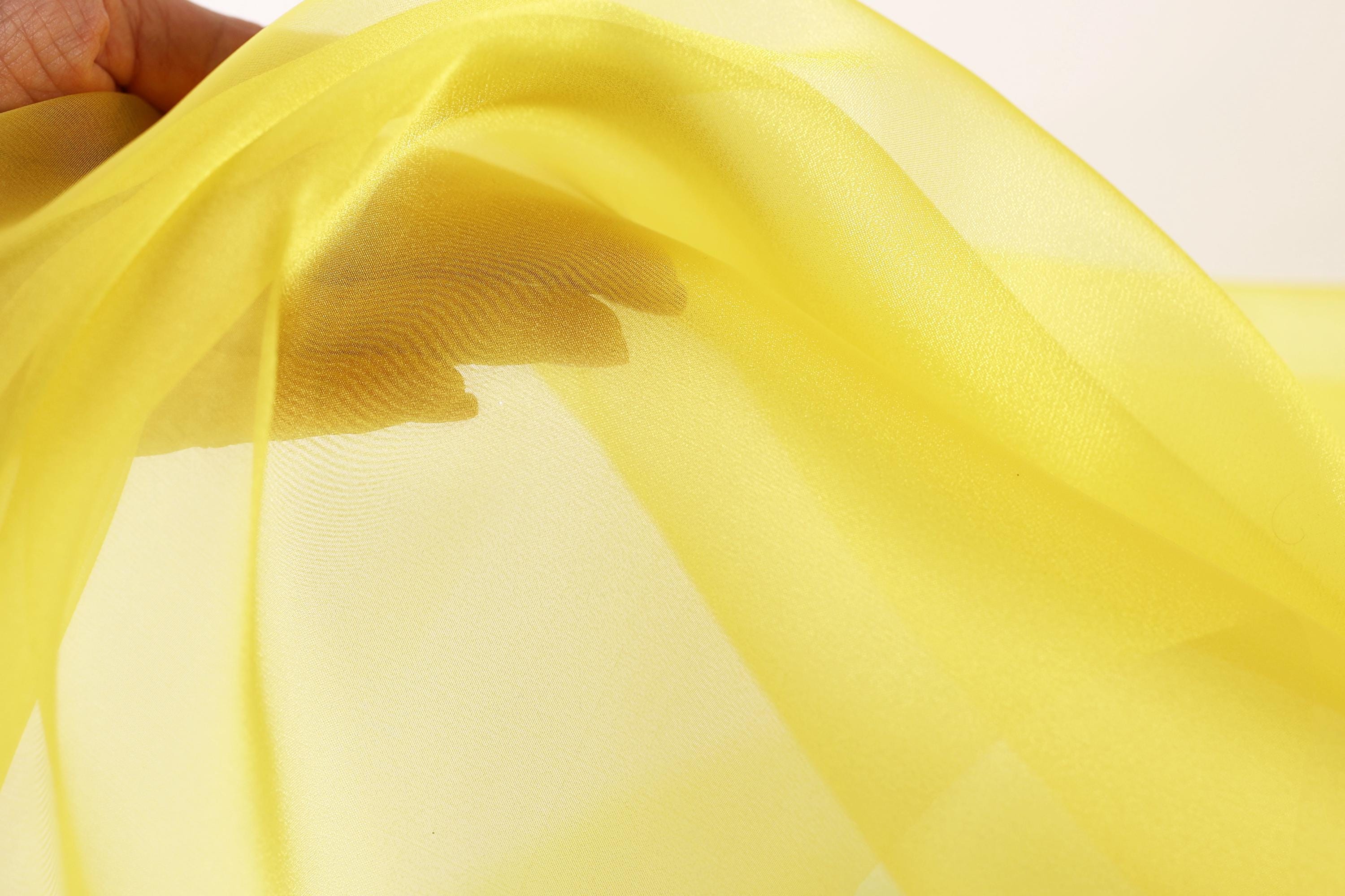 A hand is holding a yellow fabric, which appears to be a sheer material. The fabric is being held up against a white background, creating a contrast between the yellow and the white.