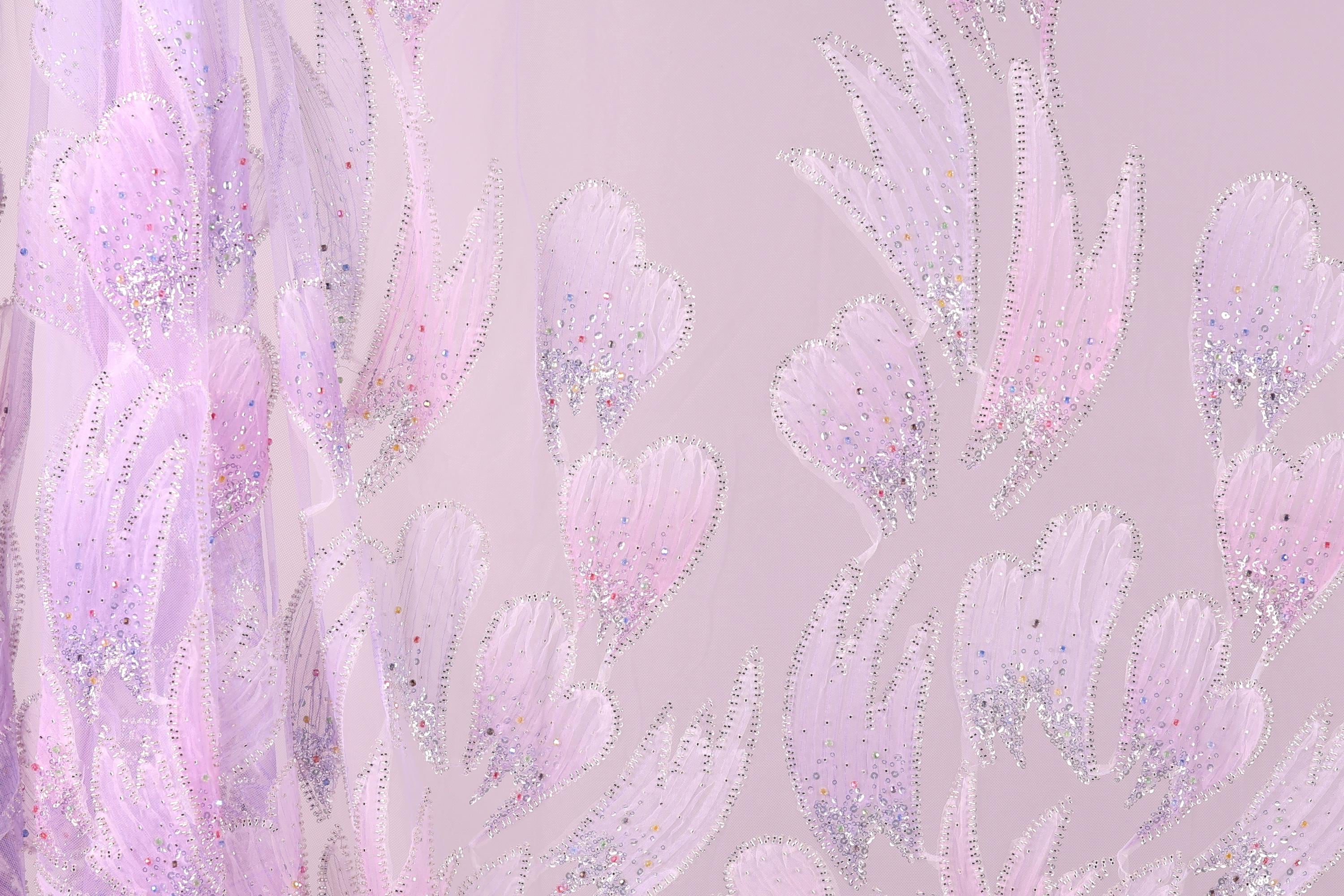 a close-up view of a fabric with a delicate, pastel-colored floral pattern. The fabric appears to be made of a sheer material, with a pink and purple color scheme. The pattern consists of numerous small, intricately designed flowers or leaves, each adorned with tiny glitter or sequin accents. The overall appearance of the fabric is soft and delicate, with a subtle, elegant aesthetic.