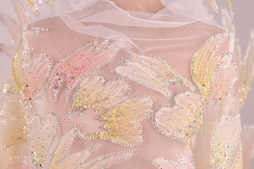 a close-up view of a delicate, intricately embroidered fabric with a floral pattern. The fabric is adorned with numerous small, sparkling beads that add a touch of elegance and sparkle to the design. The close-up perspective allows for a detailed examination of the fabric's texture and the intricate details of the embroidery.