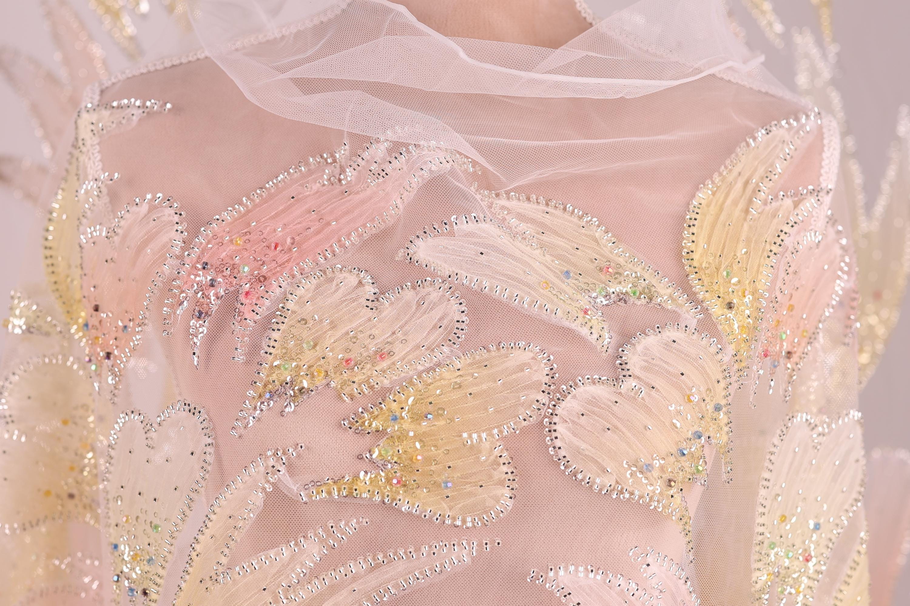 a close-up view of a delicate, intricately embroidered fabric with a floral pattern. The fabric is adorned with numerous small, sparkling beads that add a touch of elegance and sparkle to the design. The close-up perspective allows for a detailed examination of the fabric's texture and the intricate details of the embroidery.