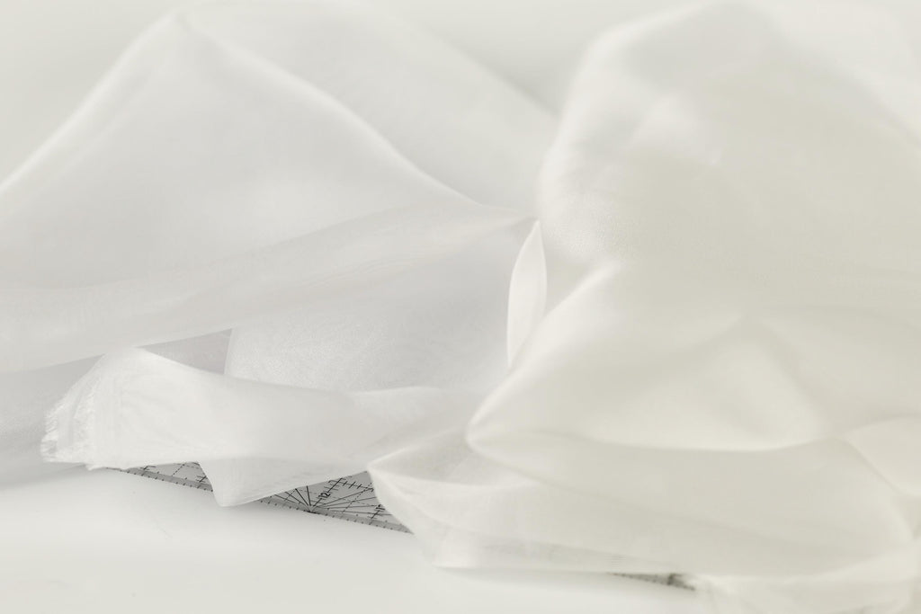 The image is a close-up of a white fabric, possibly a sheer or sheer-like material, with a subtle pattern or design visible on the surface.