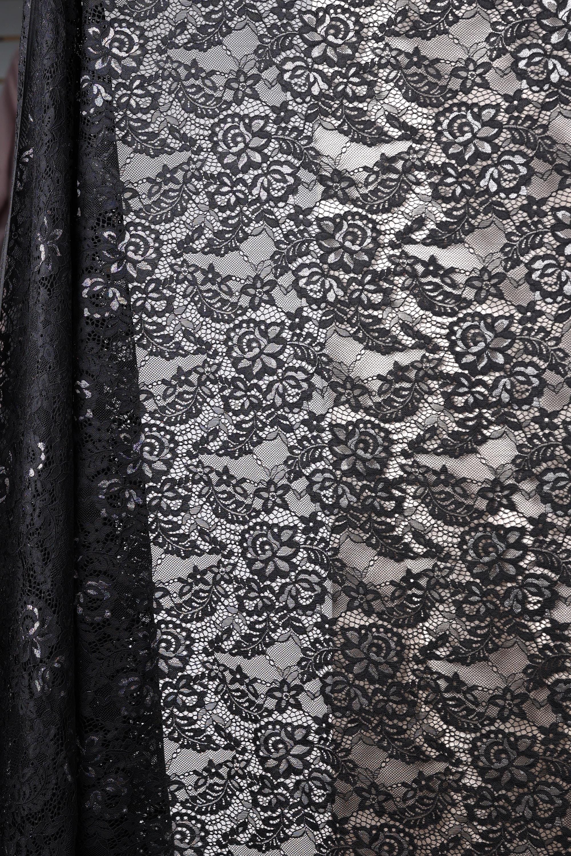 a close-up view of a black and white lace fabric with a floral pattern. The fabric appears to be a sheer lace material, with a black background that contrasts with the white lace. The lace has a delicate and intricate design, with a repeating pattern of flowers and leaves. The fabric is folded and draped, creating a visually appealing and elegant appearance.