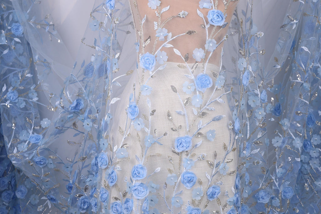a close-up view of a delicate, blue lace dress with intricate floral embroidery. The dress is adorned with numerous blue flowers and leaves, creating a beautiful and elegant appearance. The fabric appears to be sheer, allowing the light to pass through and highlighting the delicate details of the lace. The dress is the central focus of the image, with the background blurred to draw attention to the intricate details of the lace and floral patterns.
