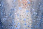 a close-up view of a delicate, blue lace dress with intricate floral embroidery. The dress is adorned with numerous blue flowers and leaves, creating a beautiful and elegant appearance. The fabric appears to be sheer, allowing the light to pass through and highlighting the delicate details of the lace. The dress is the central focus of the image, with the background blurred to draw attention to the intricate details of the lace and floral patterns.