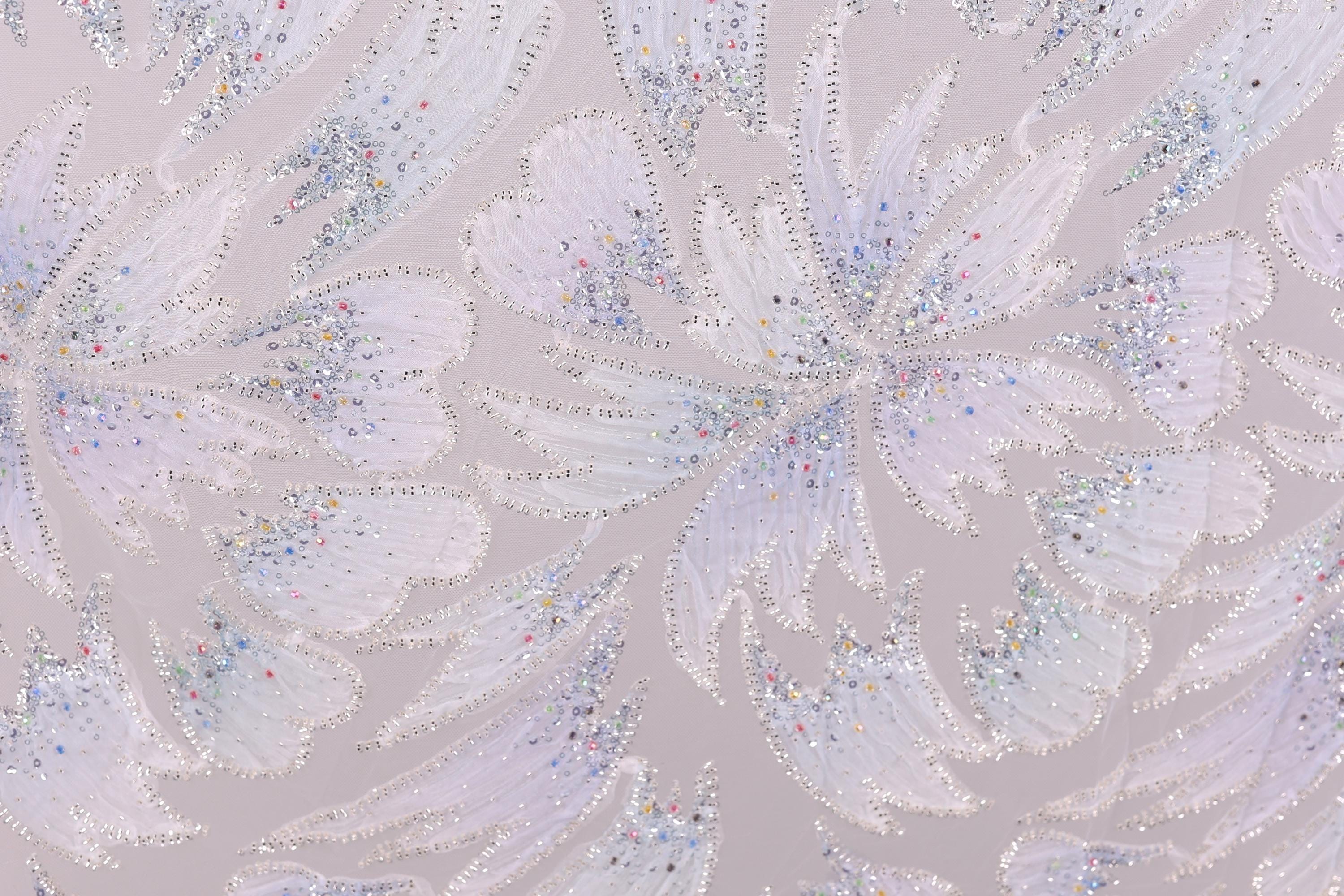 a close-up view of a patterned fabric with a repeating design of white and silver glittering flowers or leaves. The pattern is intricate and detailed, with the glittering elements creating a visually striking contrast against the fabric's texture.
