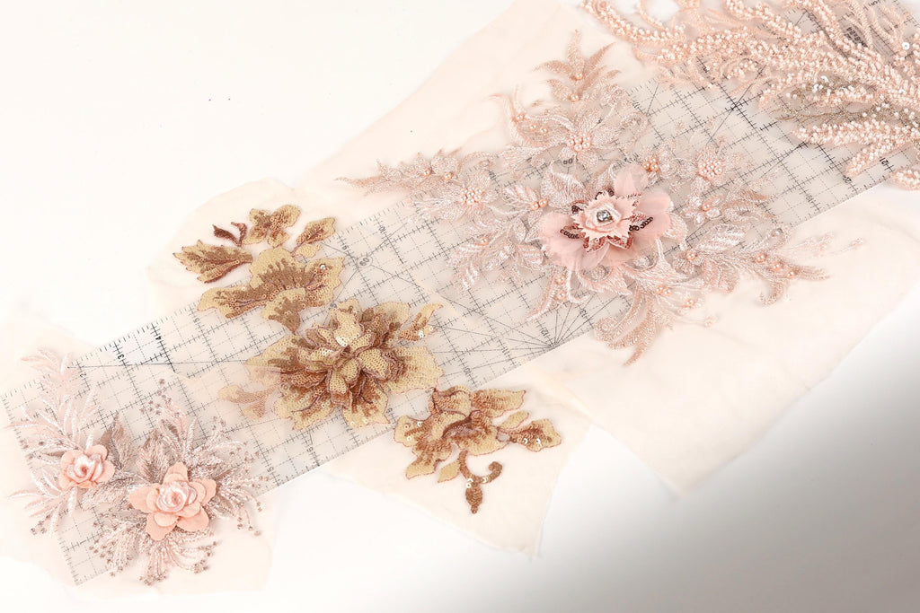a collection of delicate, intricate floral embroidery pieces, including a large pink flower with a gemstone at the center. The embroidery appears to be made from a light-colored fabric, possibly lace or chiffon, and is displayed on a white background.