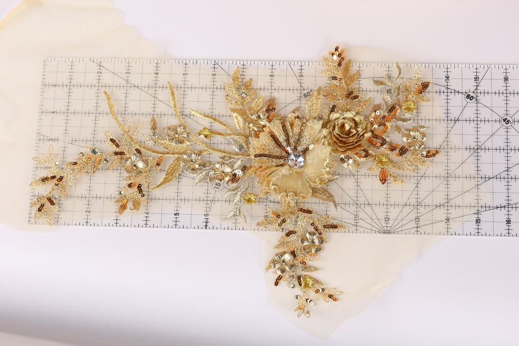 a decorative piece of gold embroidery with a flower and leaves, placed on a ruler for scale.