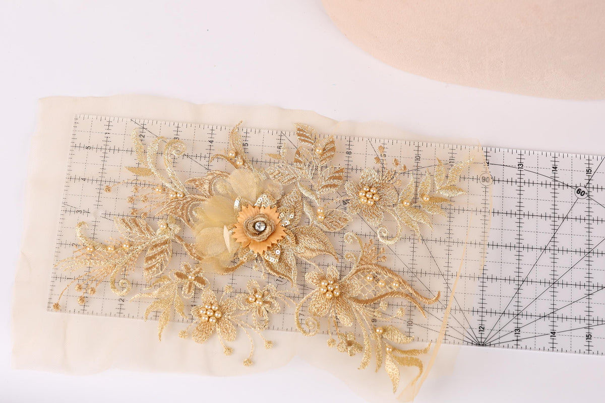 a delicate piece of embroidered fabric adorned with intricate gold floral designs. The fabric is displayed on a ruler, which is placed on a white surface. The ruler is marked with measurements, indicating that the fabric may be used for a sewing or crafting project.