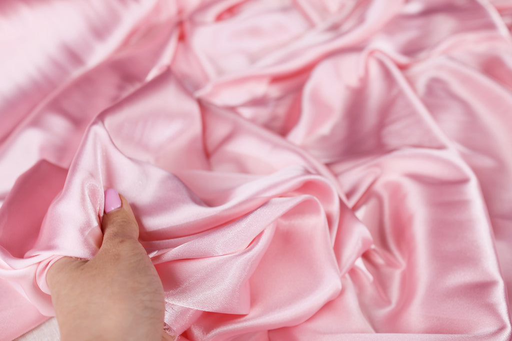 A hand is holding a piece of pink satin fabric, which appears to be soft and smooth.
