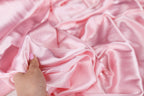 A hand is holding a piece of pink satin fabric, which appears to be soft and smooth.