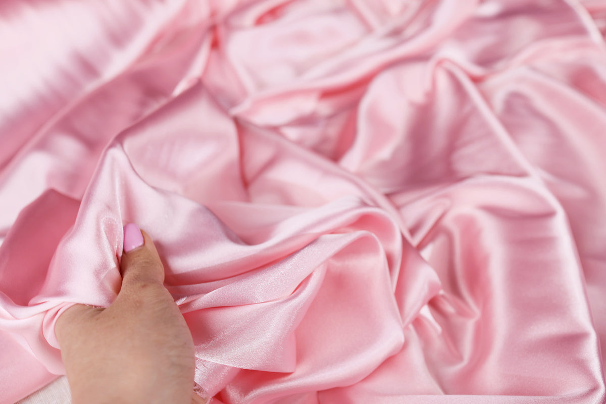 A hand is holding a piece of pink satin fabric, which appears to be soft and smooth.