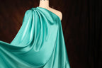 A mannequin wearing a flowing, turquoise satin dress with a draped, one-shoulder design.