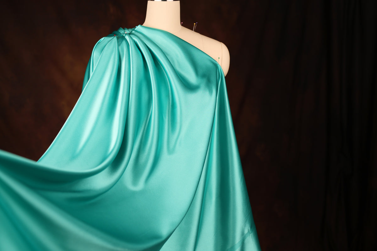 A mannequin wearing a flowing, turquoise satin dress with a draped, one-shoulder design.