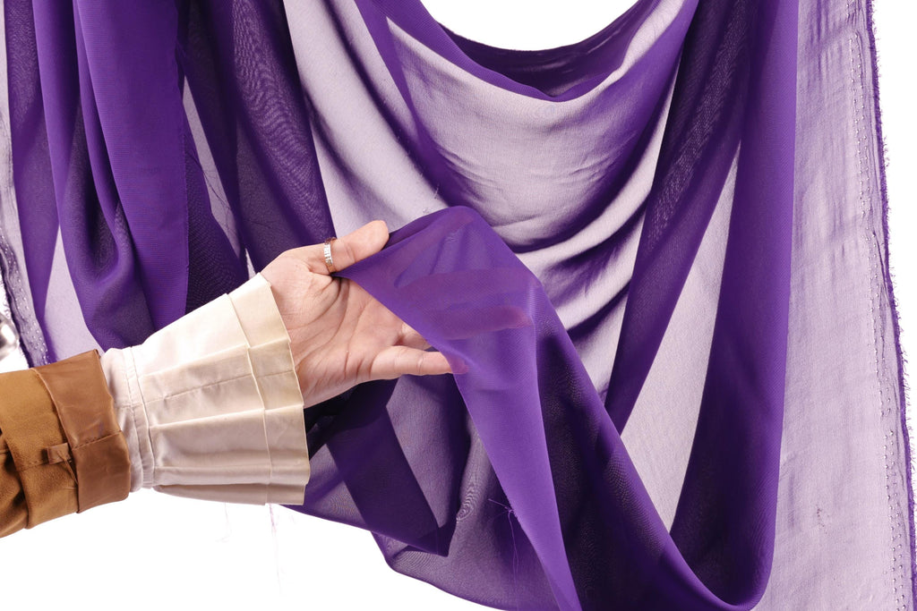 A hand is holding up a purple fabric, which appears to be a scarf or garment, against a white background.