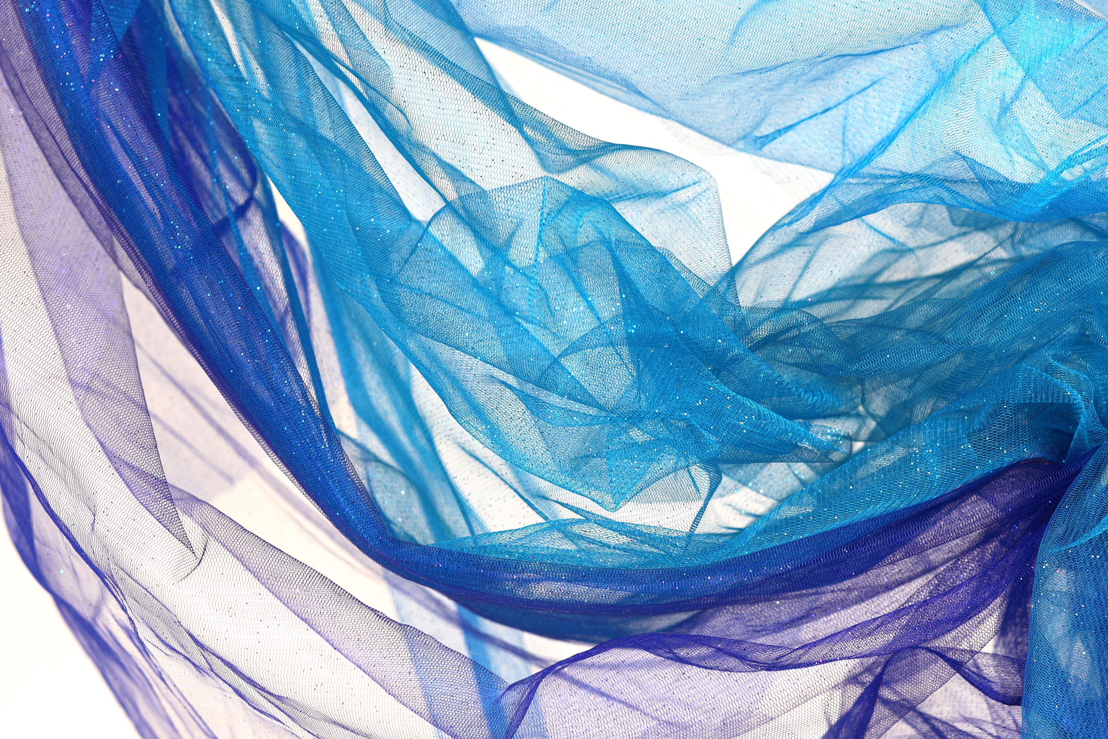 The image is a close-up of a piece of blue and purple fabric, with the fabric appearing to be made of sheer material. The fabric has a flowing, draped appearance, and the colors are vibrant and eye-catching.