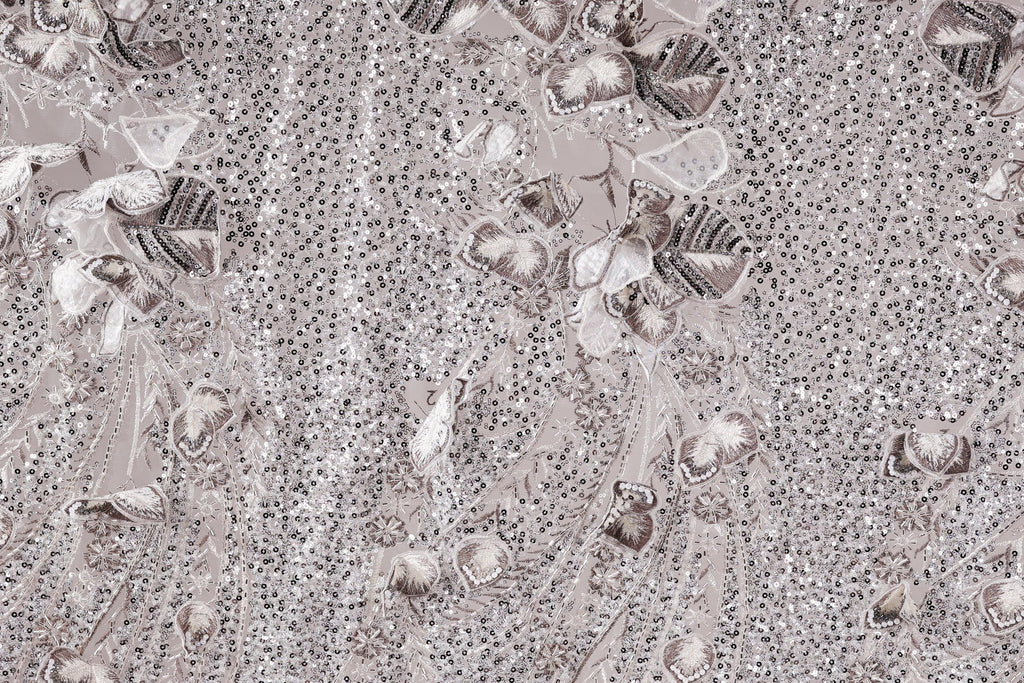 The image is a close-up of a fabric with a repeating pattern of small, intricate designs in shades of gray and white. The designs include various shapes and patterns, such as flowers, leaves, and other organic forms, creating a visually striking and detailed appearance.