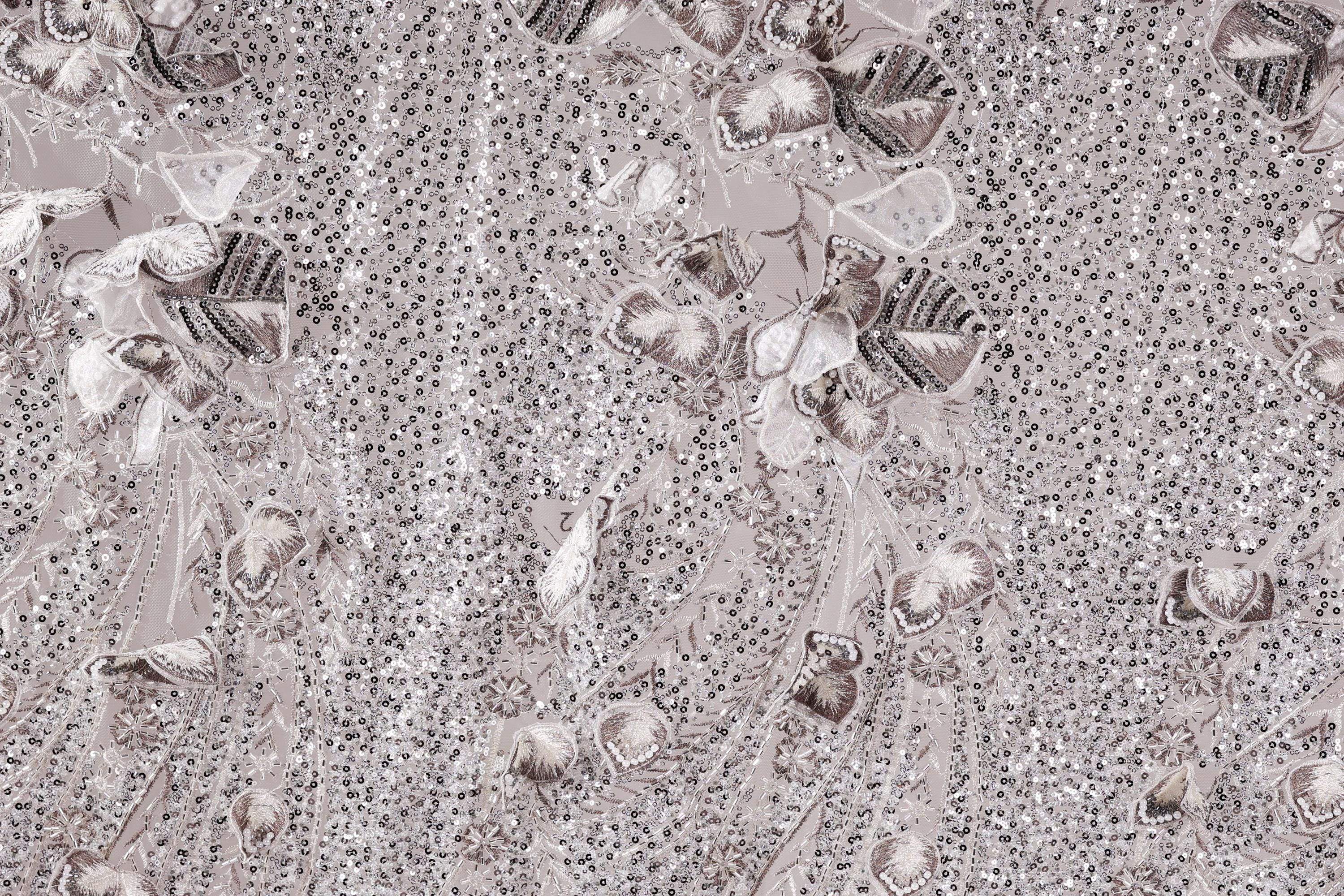 The image is a close-up of a fabric with a repeating pattern of small, intricate designs in shades of gray and white. The designs include various shapes and patterns, such as flowers, leaves, and other organic forms, creating a visually striking and detailed appearance.
