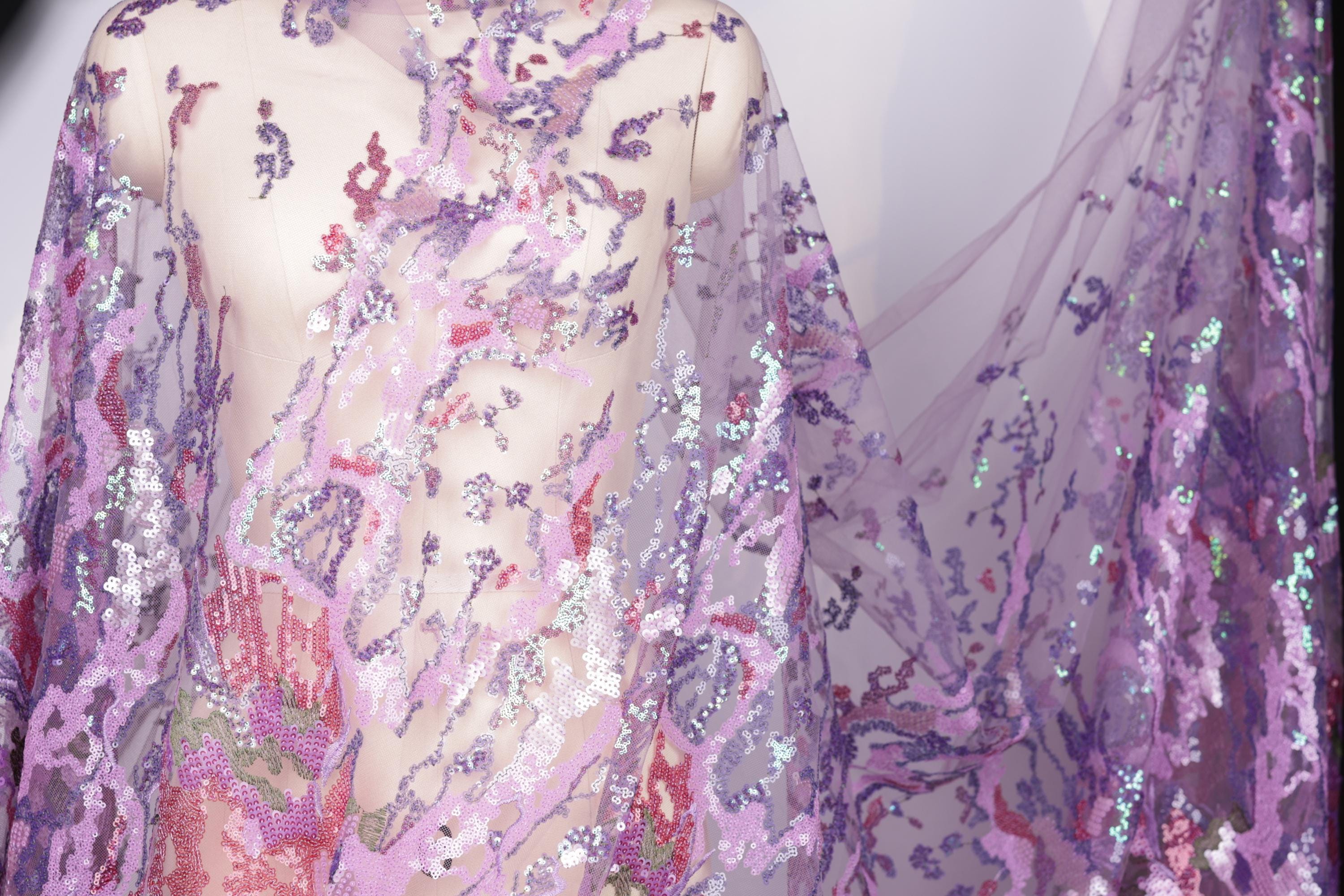a mannequin wearing a sheer, sequined fabric with a vibrant purple and pink color scheme. The fabric appears to be a sheer, lace-like material with a floral pattern, and it is draped over the mannequin's body. The mannequin is positioned in a way that highlights the intricate details and patterns of the fabric.